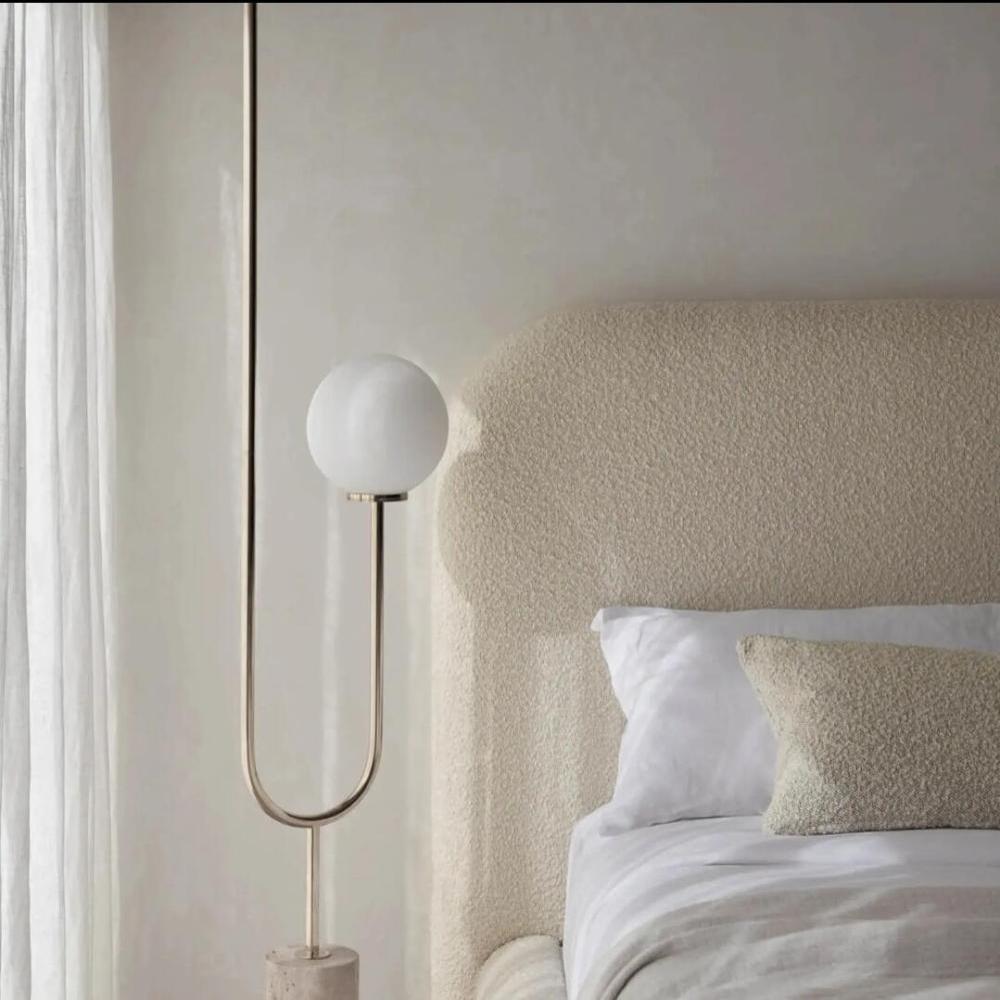 The image highlights a serene bedroom corner featuring a Teddy Boucle Bed with a curved beige upholstered headboard. A modern floor lamp with a curved metallic stand and frosted white bulb adds elegance. Soft white bedding is complemented by a textured beige lumbar pillow, while sheer curtains gently filter natural light, enhancing the room’s calm atmosphere.