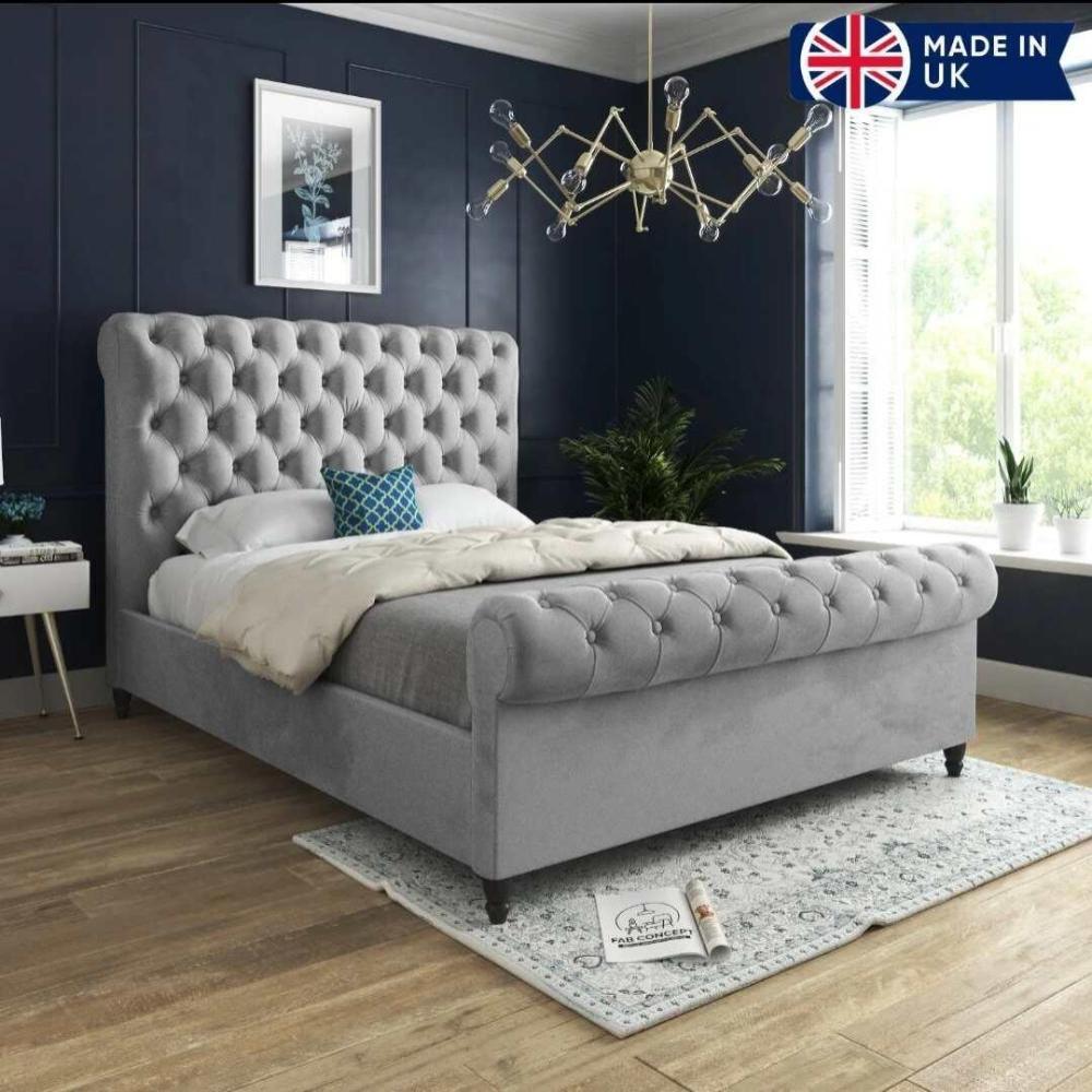 A chic bedroom features a luxurious tufted Toronto Scroll Bed in grey with a high upholstered headboard. It's styled with white pillows, a cream duvet, and a navy blue geometric pillow. Deep blue walls and a modern light fixture enhance the ambiance. Large windows bring in natural light, while a white nightstand holds a book. A patterned rug covers the wooden floor, and potted plants add greenery. A "MADE IN UK" graphic appears in the top right corner.