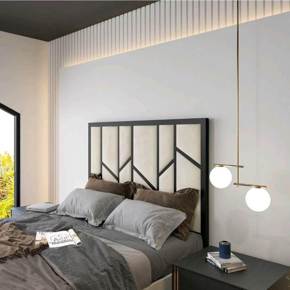 The image features a modern bedroom centered around a Zig Zag TV bed with a geometric black-patterned headboard on a light beige background. The bed is layered with gray sheets and earthy-toned pillows. To the right, a dark nightstand holds stacked books and a blue container, topped by a light fixture with two round bulbs on a golden rod.
The room is painted white with vertical paneling at the ceiling. A large window on the left fills the space with natural light and reveals greenery outside.