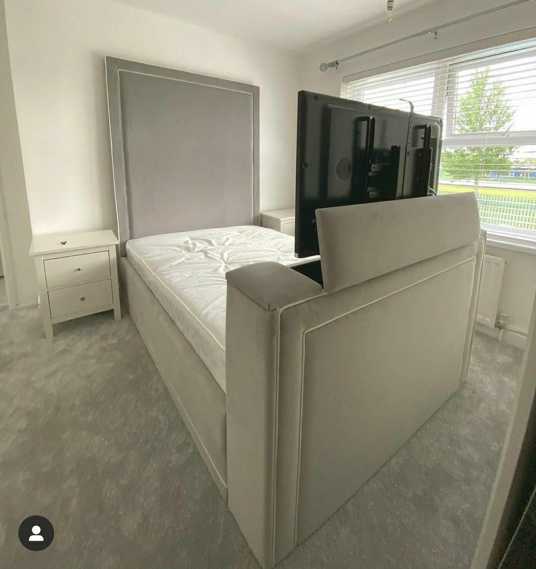 A bedroom with a large beige tufted Tv bed, white nightstand, and chandelier. Includes a wall clock, curtains, and woven baskets.