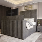 A modern grey 2025 Belgravia TV Bed with a tall, fluted headboard and footboard, featuring a large pop-up television, in a bedroom.