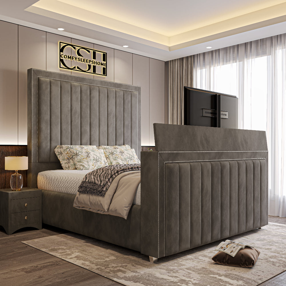 A lavish grey upholstered 2025 The Zenith TV Bed with vertical tufting, silver trim, and a pop-up TV, in a contemporary bedroom.