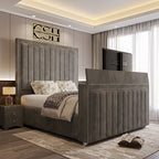 A lavish grey upholstered 2025 The Zenith TV Bed with vertical tufting, silver trim, and a pop-up TV, in a contemporary bedroom.