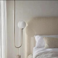 The image highlights a serene bedroom corner featuring a Teddy Boucle Bed with a curved beige upholstered headboard. A modern floor lamp with a curved metallic stand and frosted white bulb adds elegance. Soft white bedding is complemented by a textured beige lumbar pillow, while sheer curtains gently filter natural light, enhancing the room’s calm atmosphere.