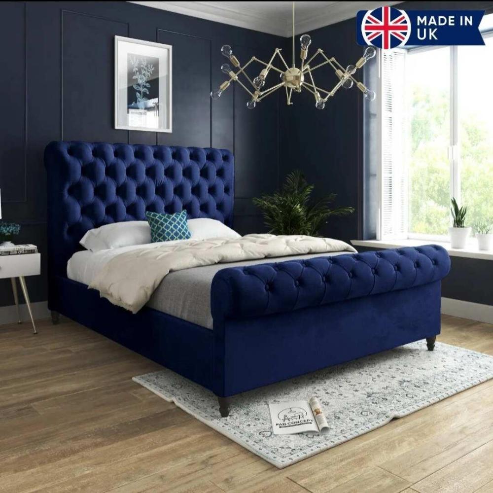 The image depicts a stylish bedroom featuring a blue Toronto Scroll Bed with an upholstered headboard. It’s dressed in white and gray bedding with a blue geometric pillow, situated on a light-patterned rug over wooden floors. An exposed bulb chandelier hangs above dark blue walls with botanical artwork. A large window on the right lets in natural light, flanked by potted plants. A small white side table is on the left, and a banner in the upper right corner displays the Union Jack with "MADE IN UK."
