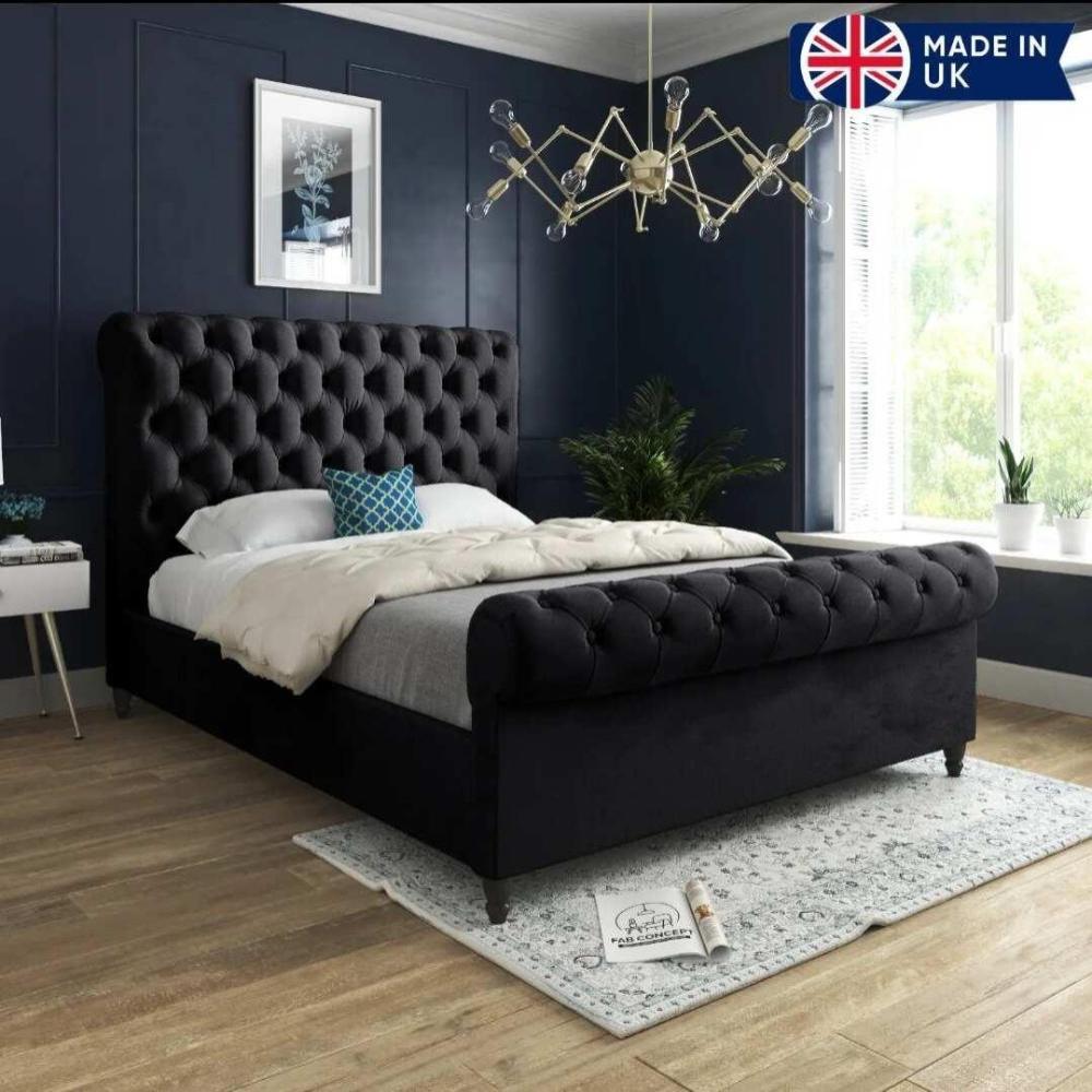 The image features a stylish bedroom with a black Toronto Scroll Bed and a high tufted headboard, dressed in a white duvet, gray throw blanket, and a blue geometric pillow. Dark blue paneled walls and an ornate chandelier enhance the elegance. A side table holds books, and a window lets in natural light, illuminating green potted plants. A light floral-patterned rug is under the bed, with a UK flag icon in the top right corner reading "MADE IN UK."
