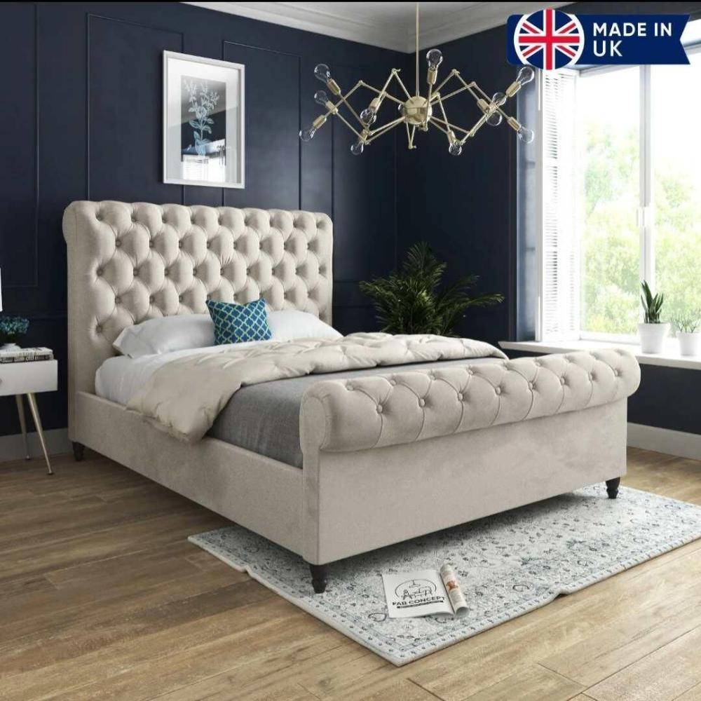 A stylish bedroom featuring the luxurious Toronto Scroll Bed with a high tufted headboard and footboard in light beige. Dressed in a cream duvet, gray blanket, and a blue and white geometric pillow, it rests on a patterned rug over warm wooden floors. A modern metallic chandelier hangs above dark blue walls, while potted plants add freshness. A "MADE IN UK" logo with the UK flag appears in the top right corner.
 Main image