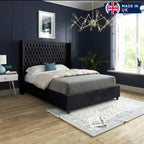 The image showcases a modern bedroom featuring a Victoria Winged Imperial Bed with a black tufted headboard, gray bedspread, white pillows, and a blue patterned cushion. The deep navy walls and gold geometric chandelier create a luxurious ambiance. A white side table with a blue lamp is positioned beside the bed. A tall indoor plant by a large window with white blinds allows natural light in. The light wooden floor is covered with a pale patterned rug. 