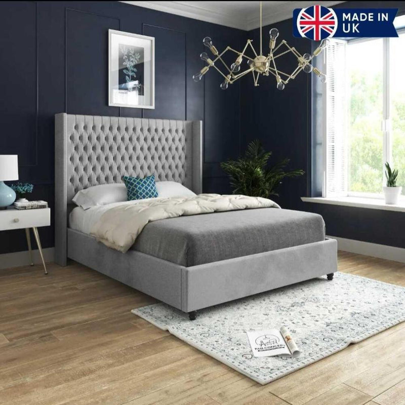 The image showcases a modern bedroom centered around a Victoria Winged Imperial Bed with a plush light gray tufted headboard. The bed features a gray comforter, white duvet, and a decorative blue pillow. A white bedside table with a blue vase and lamp sits beside it against a dark blue wall adorned with elegant molding and a plant print. A large window floods the room with natural light, highlighting a leafy green plant nearby. 