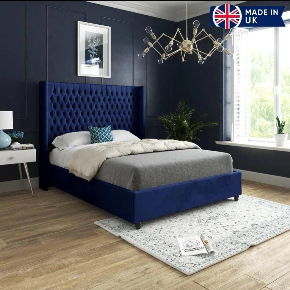 A modern bedroom showcasing a Victoria Winged Imperial Bed with a dark blue tufted headboard, gray bedspread, and white pillows. The space features a stylish chandelier and light wooden flooring.