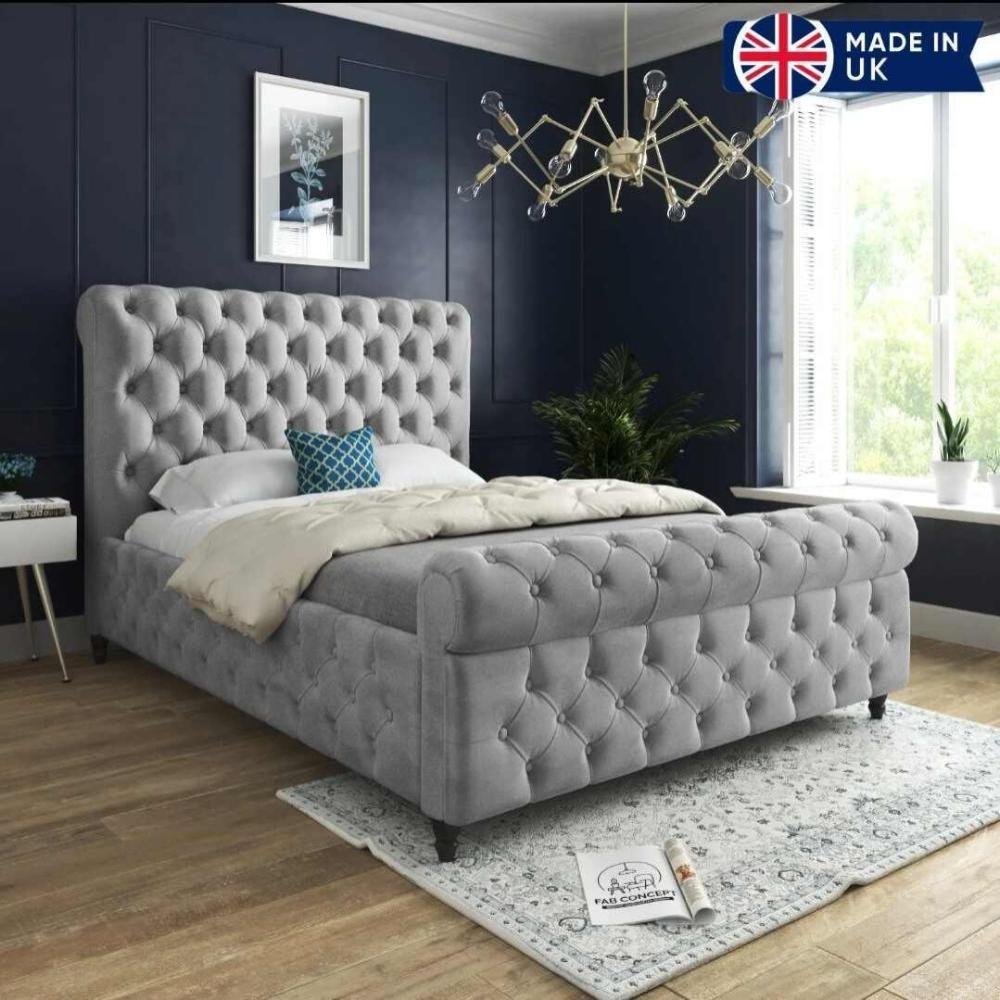 The image features a Winchester Scroll Bed with a tall, light gray tufted headboard, white bedding, and a teal decorative pillow. Dark blue walls contrast with a light wood floor and rug. A modern chandelier hangs above, and natural light brightens the room through a large window, with a potted plant nearby.