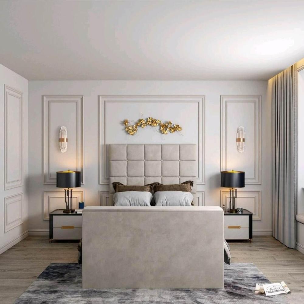 The image showcases a modern bedroom with a Windsor TV bed featuring a cream, square-patterned headboard. Gray and brown pillows complement the bed, flanked by nightstands with navy lamps and gold accents. A gold leaf wall accent decorates above, with white sconces on either side. The room has neutral walls, light wood flooring, and a gray abstract rug, with a small white cushion on the floor.