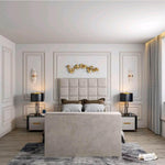 The image showcases a modern bedroom with a Windsor TV bed featuring a cream, square-patterned headboard. Gray and brown pillows complement the bed, flanked by nightstands with navy lamps and gold accents. A gold leaf wall accent decorates above, with white sconces on either side. The room has neutral walls, light wood flooring, and a gray abstract rug, with a small white cushion on the floor.