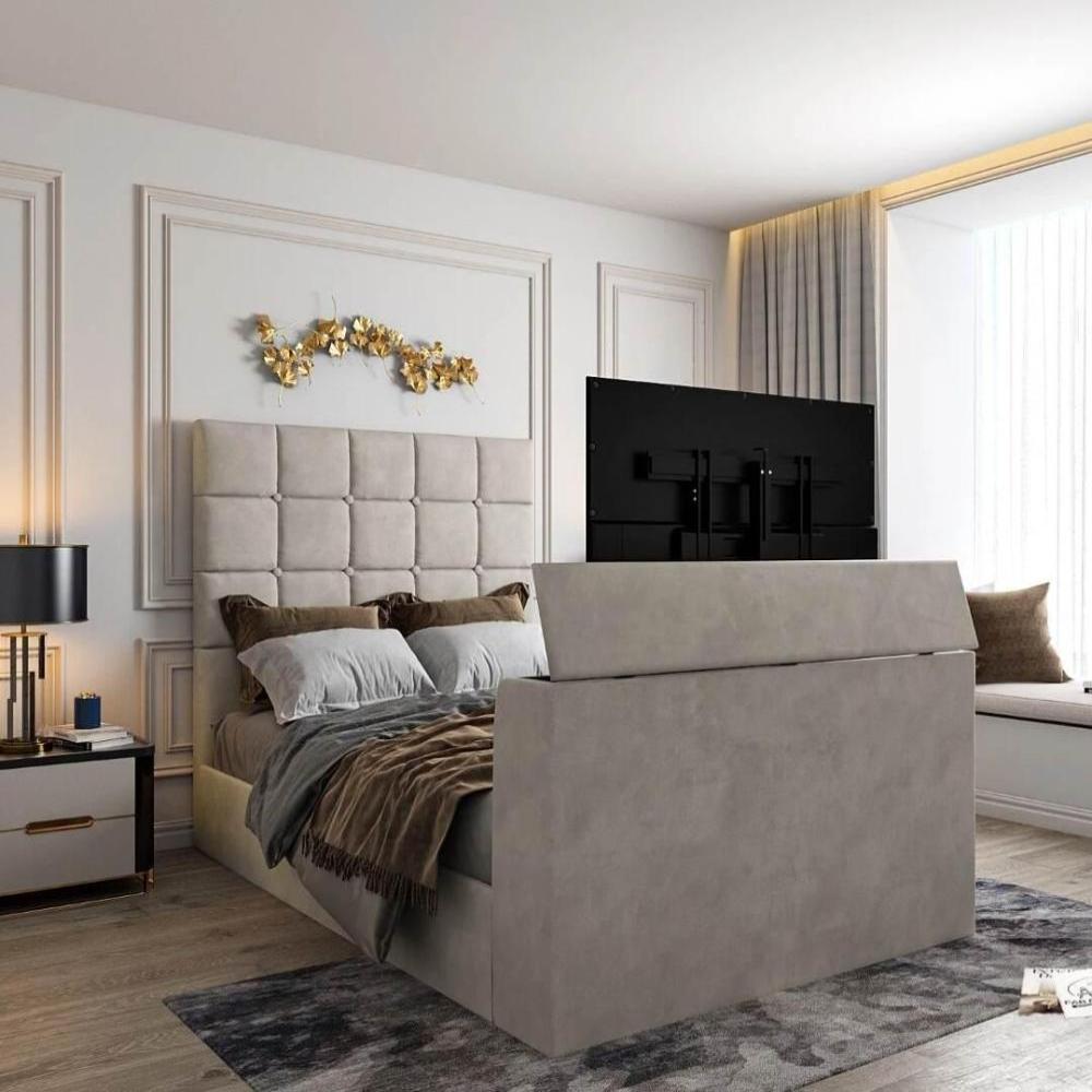 The image features a Windsor TV bed with a beige tufted headboard, gray and brown pillows, and a hidden TV at the footboard. Gray curtains frame a large window beside a cushioned seating area. A black lamp with a gold base sits on the nightstand. Above the bed, a gold leaf wall accent is framed by white moldings. Main image