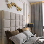 The image showcases an elegant bedroom featuring a Windsor TV bed with a tufted light gray headboard in square patterns. Four pillows—two dark brown and two light gray—adorn the bed. A gold abstract leaf art piece hangs above. To the right, muted gray floor-to-ceiling curtains frame a window, and a black table lamp with a gold rim sits on a dark nightstand, alongside a decorative object and a book.