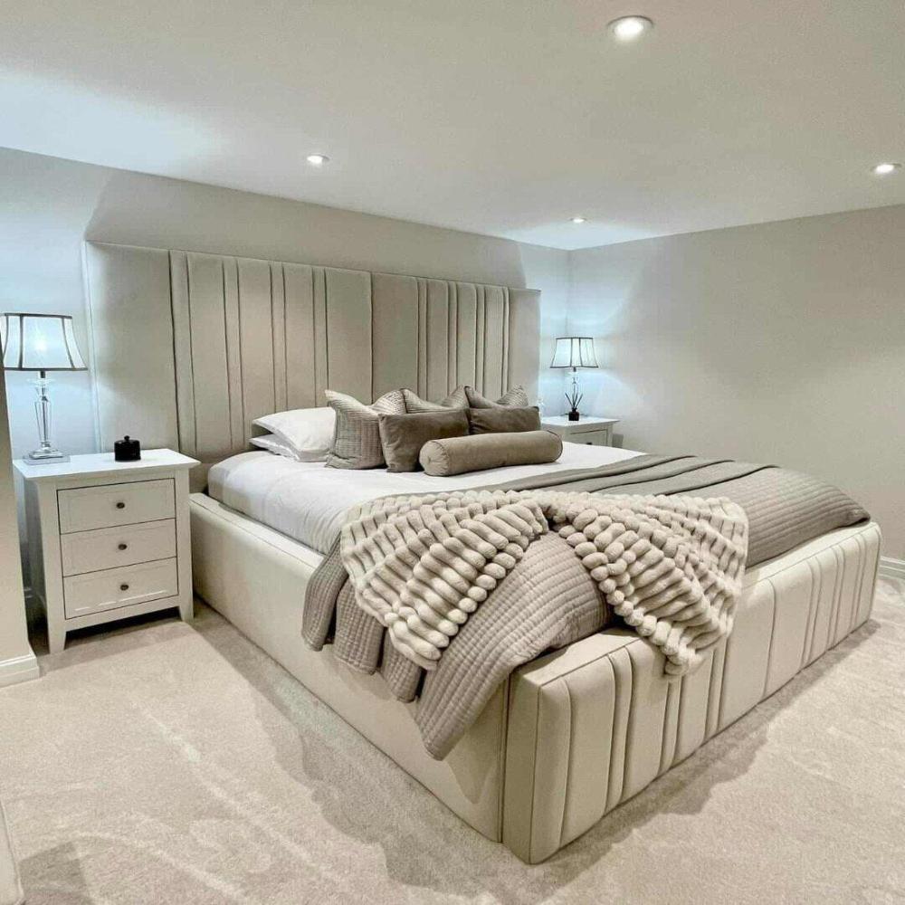 The room showcases the "Split" Bed with a soft cream upholstered headboard and white linens accented by beige and taupe pillows. A textured throw adds warmth at the foot. White bedside tables with matching lamps and recessed ceiling lights enhance the minimalist, serene vibe. A plush, light-colored carpet completes the cozy look.